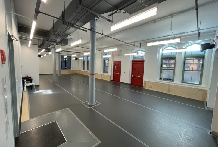 A dance studio with a grey marley floor, windows, and red doors. There are three grey columns distributed through two thirds of the space.