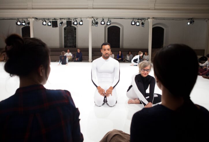 2 performers sit on a white marly floor in all white and all black unitards. The silhouette of the back 2 people's heads in the foreground and groups of people in the background behind the seated dancers.