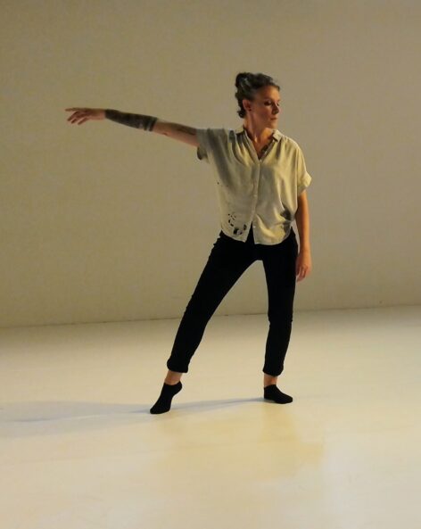 In a white space with warm lighting a woman stand with all her weight on her left leg. She is wearing black pants and socks and a white short sleeve shirt. Her hair is pulled back and she looks to the left. Her right toes are touching the floor to the right of her center and her right arm is raised out to the side, hand relaxed. Photo by Christopher Tan.