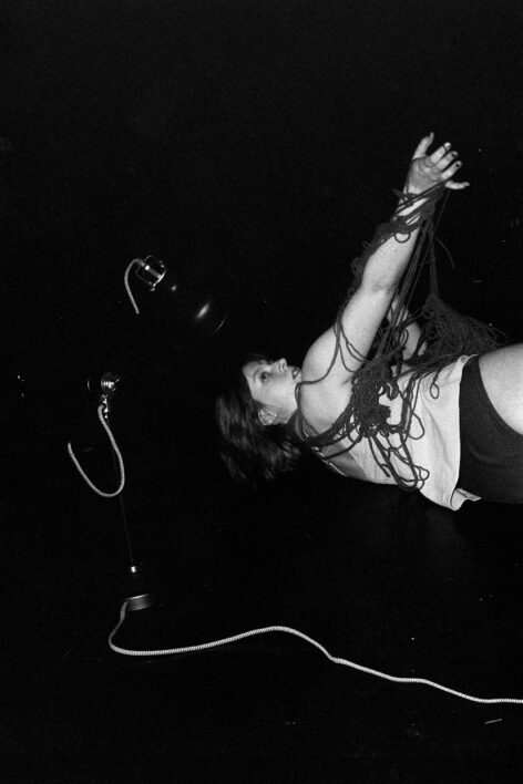 A black and white photo of Meg with a dark background. She is laying on her left side with her back to the camera, only her torso is in the image. Her right arm is extended and she is looking up and over her right shoulder. She is entangled in yarn and there are three white chords in the background. Photo by Shosh Isaacs.