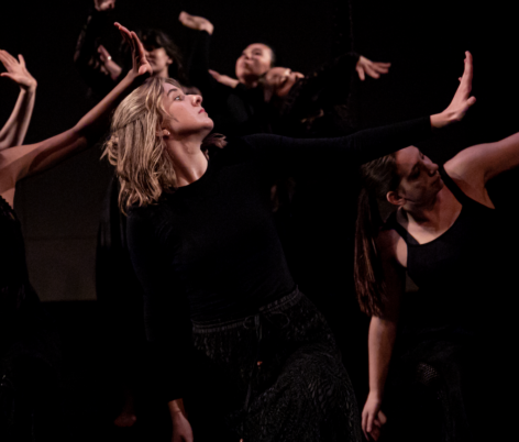 Dressed in black from the neck down, Olive is seen profile, looking out over their left outstretched fingers that reach away from them on a diagonal. They are in the forefront of a group of other dancers performing the same move, also in black.
