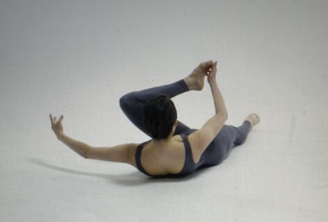 An image of a dancer, Daphne Fernburger, lying on her back and holding her foot with opposite hand. A single image from video of Spirals filmed by Rafe Scobey-Thal.