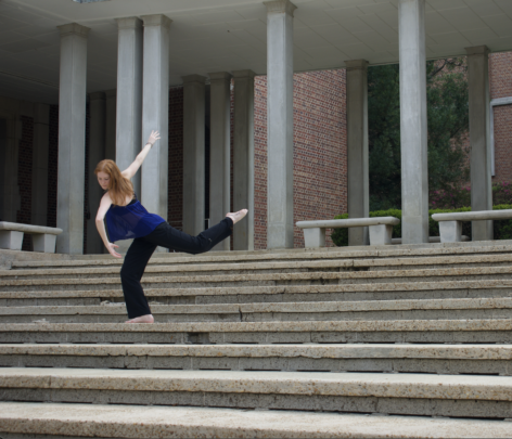 Alex is on the steps of an outdoor theater space in a blue tank top and black pants. She is extending her back leg behind her while looking over her left shoulder.