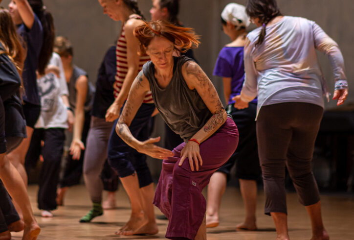 A photo from Vicky Shick's workshop for MELT 2023. Dancers move close to each other. In the center a dancer wearing purple pants stands with knees bent. One hand rests on their upper thigh while their torso spirals following the movement with their other arm forming a curve. Their short orange hair whips around their face. Photo by Rachel Keane