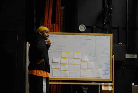 A tall man in a black hoodie and yellow bandana stands in front of a white board with small sheets of paper taped to it; His hand grazes his mouth as h looks out into a dance studio. Photo by Randy Karels.