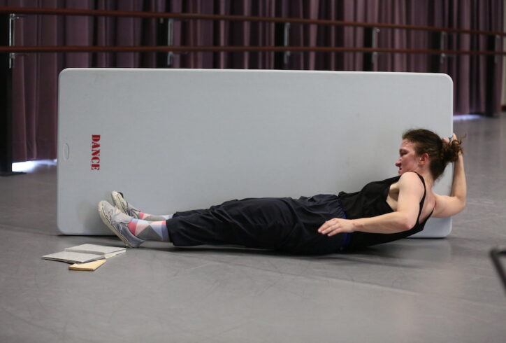 Full body image of Jeanine in black clothing with multi-color socks, books at her feet, and worn out Van sneakers, in the midst of laying down while holding onto the edge a long folding table with the word 