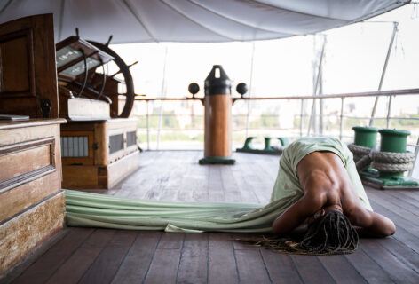 Photo by Cherylynn Tsushima. A photo of a person's exposed dark brown back on the deck of a boat laying in child's post, they wear a light green sheet and their long black and blonde braids drape over them.