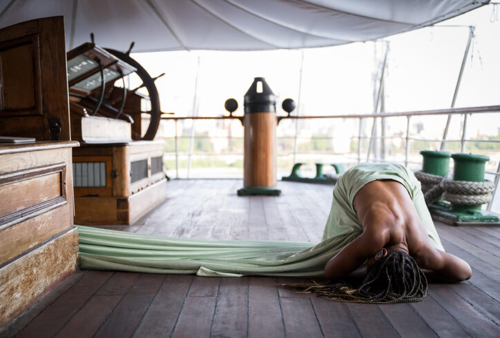 Photo by Cherylynn Tsushima. A photo of a person's exposed dark brown back on the deck of a boat laying in child's post, they wear a light green sheet and their long black and blonde braids drape over them.