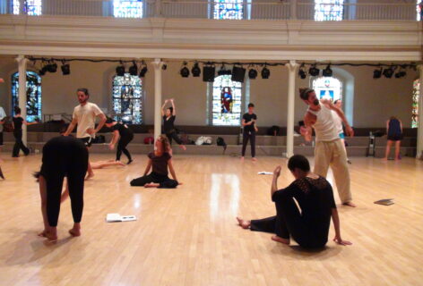 12 adult dancers are spread across the wooden floor of St. Mark's Church, with three notebooks lying on the floor as well. The dancer are white, brown, and Asian, some masculine-presenting and some feminine-presenting. Two dancers are in the midst of movement on the floor, the remainder of the dancers are in the midst of movement whilst standing. No two dancers are alike. Photo by Ashley. a friend of Movement Research.