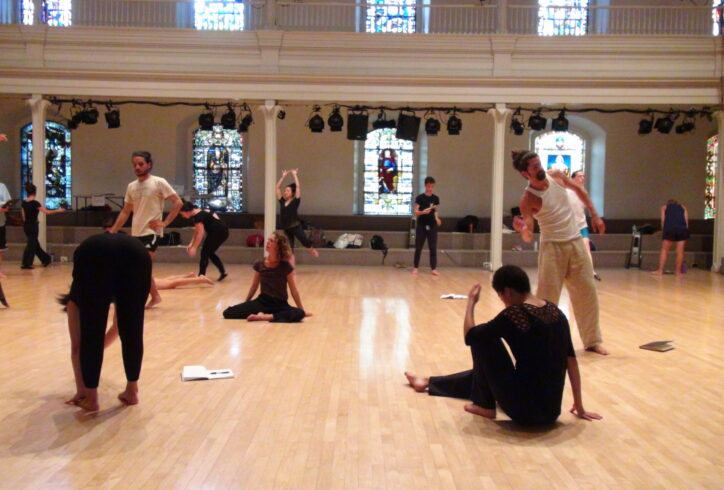 12 adult dancers are spread across the wooden floor of St. Mark's Church, with three notebooks lying on the floor as well. The dancer are white, brown, and Asian, some masculine-presenting and some feminine-presenting. Two dancers are in the midst of movement on the floor, the remainder of the dancers are in the midst of movement whilst standing. No two dancers are alike. Photo by Ashley. a friend of Movement Research.