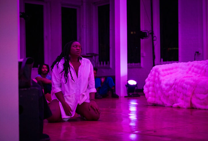 Ogemdi Ude, a dark skinned Black femme is wearing a white button down dress, her hair in long twists; She is on her knees and looking towards a fluffy white couch in a room full of people. Photo by Cherlynn Tsushima courtesy of The Kitchen.