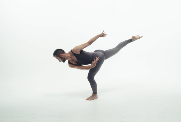 A male dancer in a grey leotard balances on one leg with one arm extended back. This is a screenshot from the video Spirals, provided by the Artist.