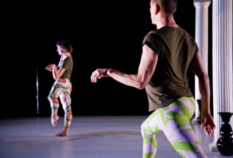 Two dancers in different positions, both wearing boldly patterned baggy-crotch tights and dark t-shirts, and both with limp wrists, hands hanging. The stage on which they are dancing also includes a microphone stand and two white columns, with black vases next to the base of each column. photo by Frank Mullaney of Johnni Durango and Neil Greenberg in Greenberg's 