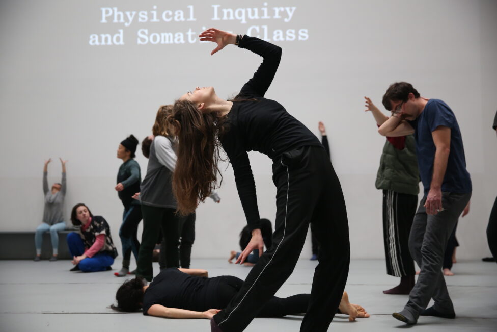 2019_Jan 2019.MR@MoMA. iele paloumpis_Jaime Ortega.Photo by Paula Court.155 Photo from iele paloumpis’s workshop for Movement Research at MoMA. Dancers in motion, a dancer in the foreground stands arching backwards. Text projected on the wall reads, Physical Inquiry and Somatics Class. Photo by Paula Court.