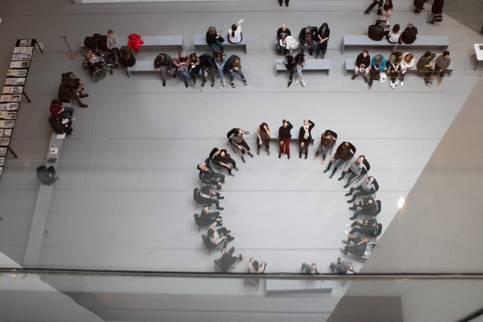 2019_MR@MoMA.2019.Jimena Paz.Photo by Andrew Kim.22 Photo from Jimena Paz’ workshop for Movement Research at MoMa. Photo taken from above. People sit in benches while a group sits in a circle on chairs with their right hand placed on their heads. Photo by Andrew Kim.