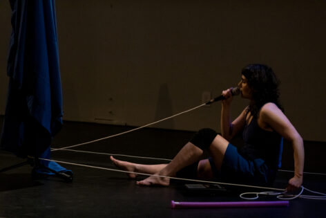 Allie, a white woman with dark hair, sits on the ground in a black box theater speaking into a microphone with a white mic cable. Photo by Michelle Smith Lewis.