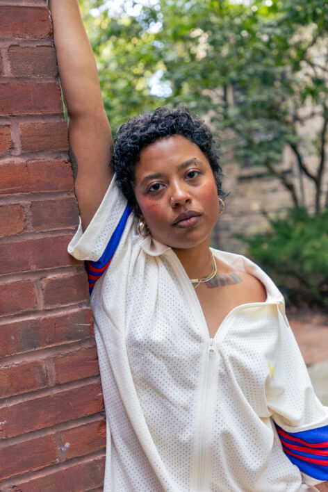 Symara Sarai a black femme leans against a brick building with one arm up and the other to her side. She looks at the camera directly, behind her is a landscape of greenery. She's wearing a white zip down shirt that's open to her sternum, the sleeves are rimmed with the colors red and blue. She's adorned in gold on her neck and ears with jewelry passed down from her ancestors. She has short dark brown curly hair and her face has a generous amount of blush to show a flushed look. Photo Credit to Whitney Brown.