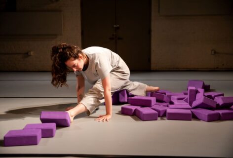 Sofia, a white brunette dressed in loose grey athletic gear, approaches a split with rounded spine. She is surrounded by purple yoga blocks. Photo by Rachel Keane.