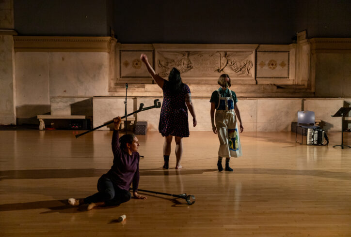 Kiera Bono, Michelle Mantione and Krista Miranda perfoming for Movement Research at the Judson Church. One dancer sits on the ground holding one crutch above their head and resting the other on the ground. Two other artists stand nearby. One waves their arm upwards. Photo by Rachel Keane.