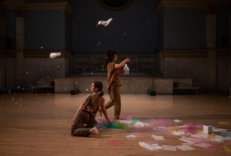 Photo from Halie Bahr presents Human Resources-HR Department performing for Movement Research at the Judson Church. One dancer kneels while another walks near them taking thin sheets of paper out of a kleenex box. There are tissues in the air and on the ground along with tulle fabric. Photo by Rachel Keane.
