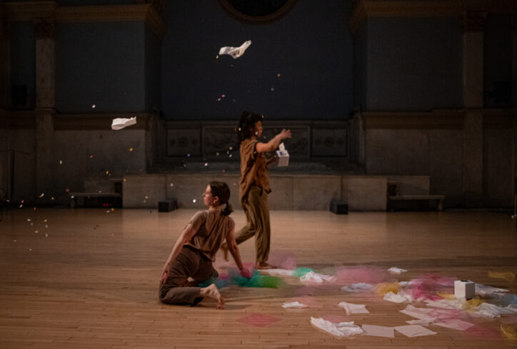 Photo from Halie Bahr presents Human Resources-HR Department performing for Movement Research at the Judson Church. One dancer kneels while another walks near them taking thin sheets of paper out of a kleenex box. There are tissues in the air and on the ground along with tulle fabric. Photo by Rachel Keane.