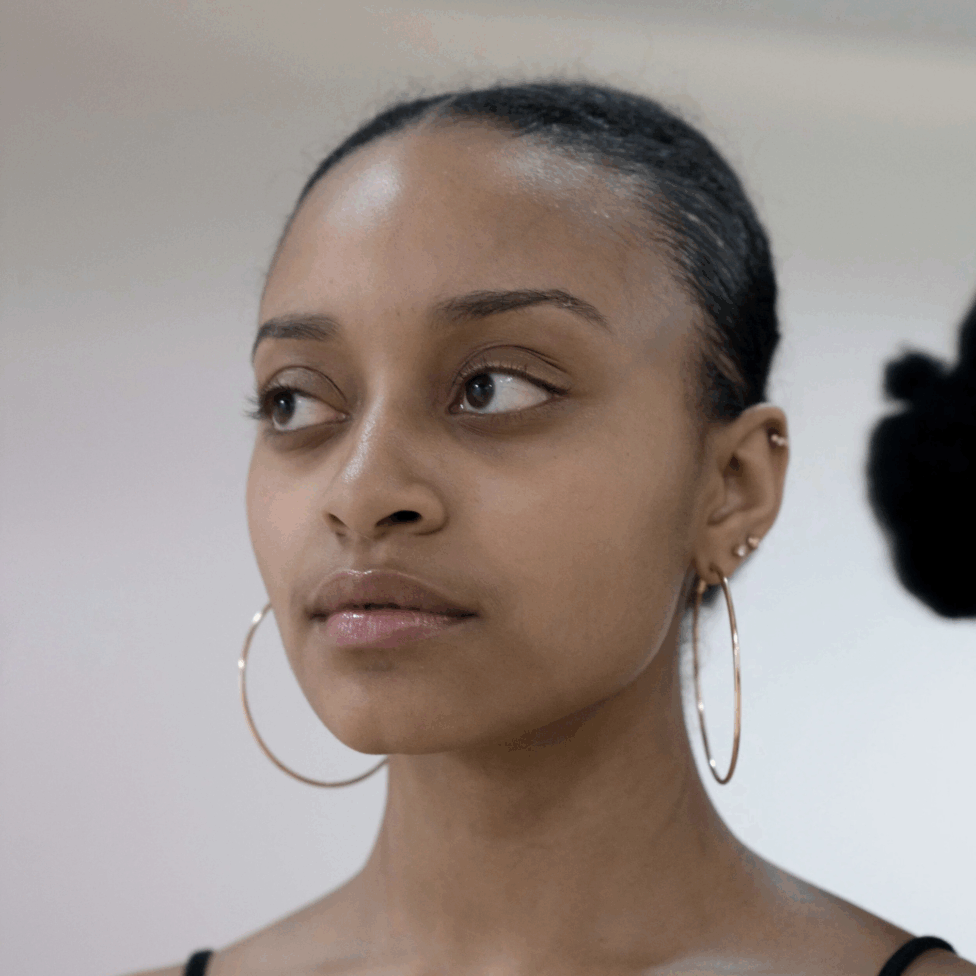 Close-up portrait of Alysia with slicked-back hair and large hoop earrings, gazing left in soft studio light.
