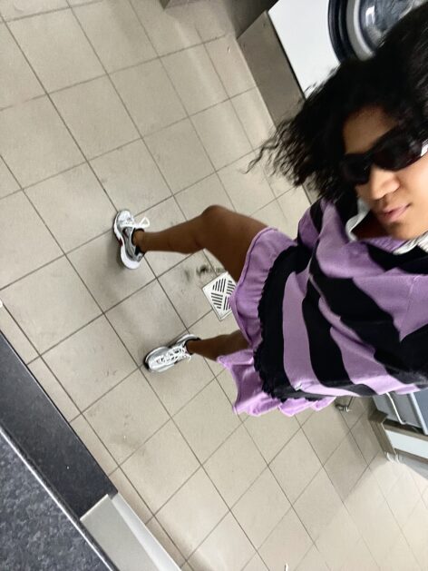 A black person in a black and purple stripped jersey sweatshirt dress framed in the right edge of the photo in a laundry room. Photo by Lydia Östberg Diakité