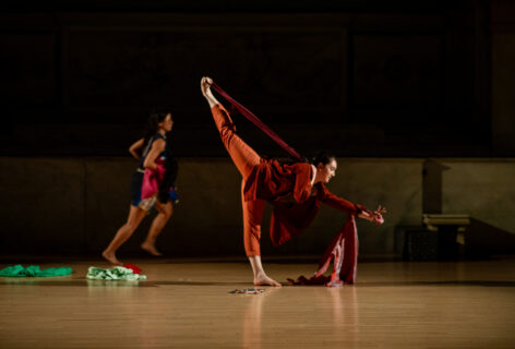 Photo from Tina Bararian's piece for Movement Research at the Judson Church. A dancer runs int he background. In the foreground a dancer stands balancing on one leg with the other reaching upwards to the ceiling. A band tied around the ankle is pulled forward. Photo by Rachel Keane.