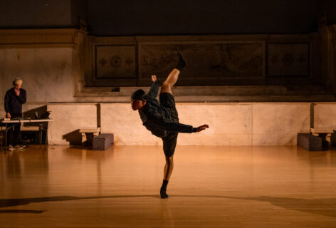 A photo of Morgan Gregory's work for Movement Research at the Judson Church. A dancer balances on one leg swinging the other leg behind and upwards while opening their hips. In a background a DJ plays music. Photo by Rachel Keane.