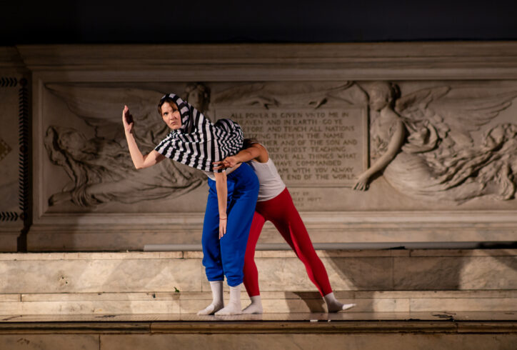 Photo from Sasha Vega's piece for Movement Research at the Judson Church. One artist stands with their shirt over their head while another stands behind them with their head inside the first artist's shirt. Photo by Rachel Keane.