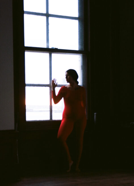 I am wearing a red one-piece standing next to a large window back-lit with overcast light shot on 35mm film. Photo by Muyassar Kurdi.