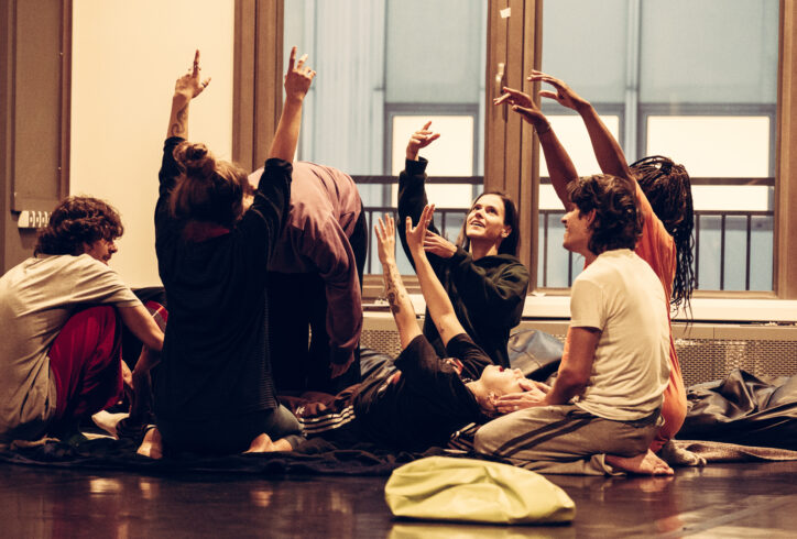 Six dancers kneeling in a circle laying hands on a dancer laying down. Three dancers have their arms raised, two have their hands on he person laying down, and one has their hand supporting the head of the one laying down. The person laying down has their mouth open, eyes closed and hands raised. The dancers are in colorful clothing and their faces are filled with elation. The light is bright in a room with a black marly dance floor.
