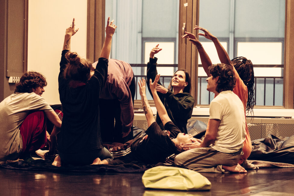 Six dancers kneeling in a circle laying hands on a dancer laying down. Three dancers have their arms raised, two have their hands on he person laying down, and one has their hand supporting the head of the one laying down. The person laying down has their mouth open, eyes closed and hands raised. The dancers are in colorful clothing and their faces are filled with elation. The light is bright in a room with a black marly dance floor.