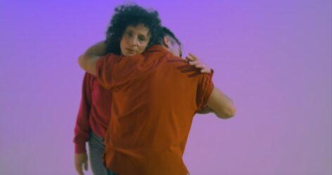 Two dancers stand, physically wrapped around each other, one with their back to the camera with medium length black hair wearing a rust colored shirt, the other facing out, with long curly hair and wearing a pink sweatshirt, with her hand on the other's shoulder. Video Still by Chico Eastridge.