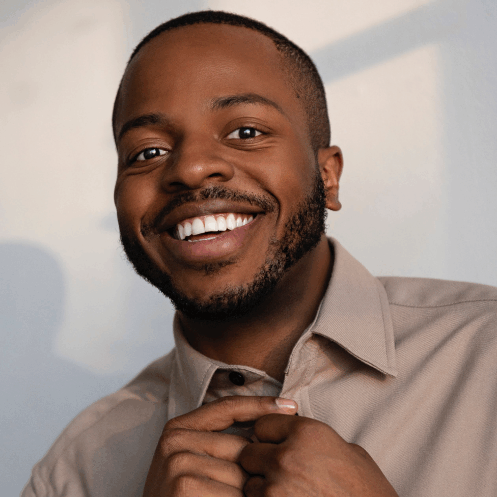 Smiling portrait of Antuan in a beige button-up, hands adjusting the collar, warm window light on a neutral wall.