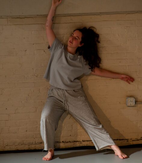 Sofia, a white femme with long brown hair, leans against a white brick wall while dressed in grey sweats. Photo by Rachel Keane.