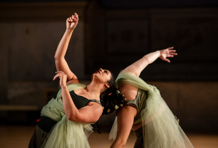 RACHEL dancers performing for Movement Research at the Judson Church. Two dancers draped in long green pieces of green tulle stand together leaning towards each other. One reaches their arm upwards gesturing. Photo by Rachel Keane.