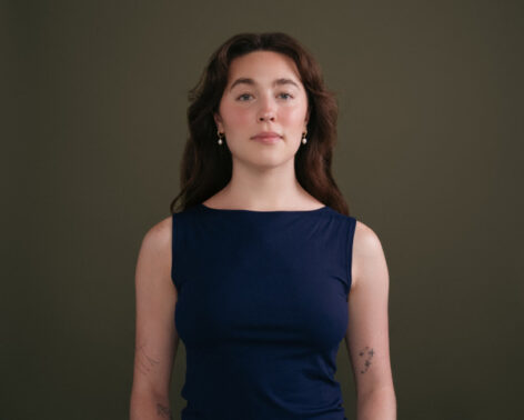 This upper body photo features an olive green background, Ally’s long, wavy brown hair, pearl earrings, and a navy blue top with a slight smile expression.