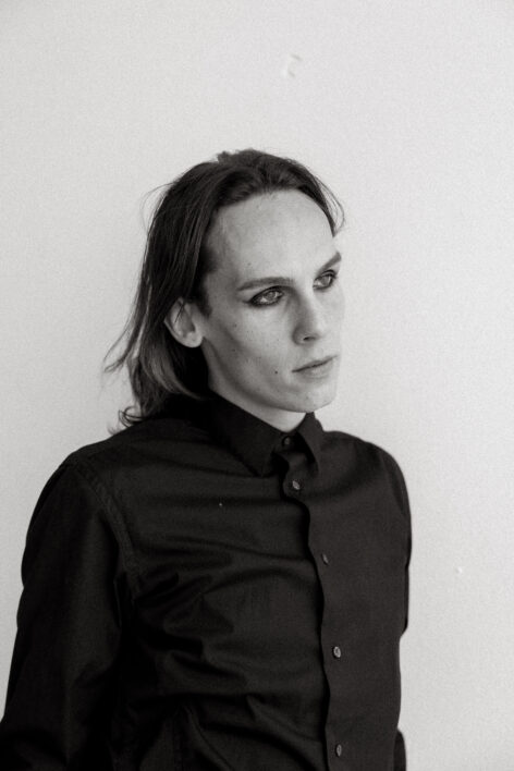 A black and white headshot of the artist. They wear a dark button down and eyeliner. Photo by Hannah Mayfield.