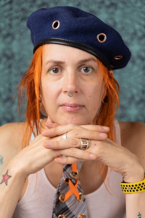 A person with orange hair, wearing a blue beret with metal grommets, a tank top and a tie, against a lime washed dark green background. PHOTO BY EXUM FOTOGRAFIE.