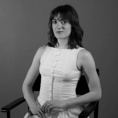 Black and white portrait of Calie. She is seated, wearing a white sleeveless button down. She is looking into the camera with a neutral expression.