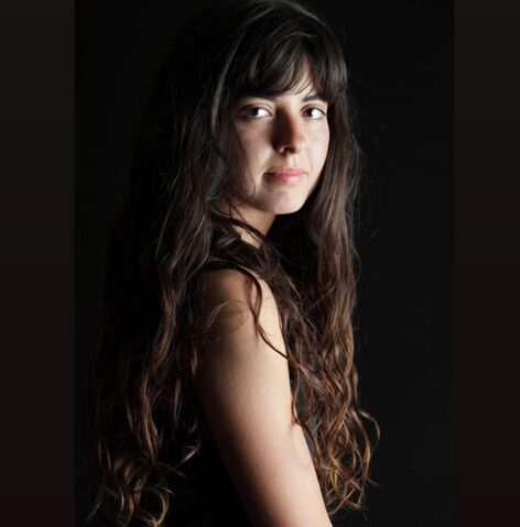 A close-up portrait of a person with long brown hair and brown eyes, fair white skin, and a subtle smile. Their head is slightly turned to the right, with their gaze gently looking over their shoulder. They wear a black shirt, and the expression is calm and soft. Photo by Hadas Danon.
