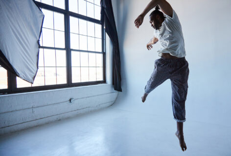 Jordan Demetrius Lloyd, a tall black man leaps in the air wearing black striped pants and a white shirt. His left leg is extended down toward the white floor while his right leg reaches toward the back corner of the room, which has a wall of windows with frosted glass. Photo by Whitney Browne.