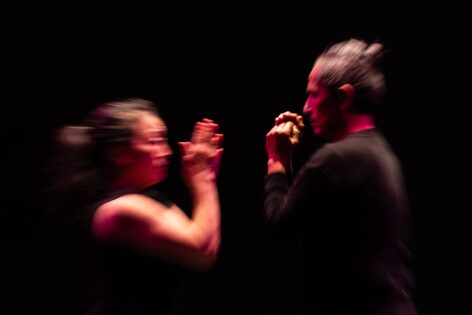 Two people face each other against a dark background with dramatic lighting. The both have brown skin and dark hair and have their hands raised in between them. The image is slightly blurred in movement. Photo by Robbie Sweeny