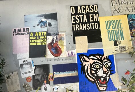 A cement wall filled with posters with different dimensions and colors, some containing words in Portuguese others in English, all related to art as resistance, collective life, trans & queer activism, and decolonial struggle.