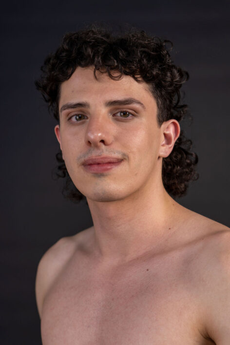 Parker Ramirez (they/them) against a black background facing directly at the camera as their torso is angled toward the left side of the picture. They are a soft masculine looking individual, they have a curly shag/mullet haircut that extends to the back of their neck and covers their forehead partially. They have a slight shadow of stubble around their lips and chin. Photo Credit is Nicholas Talamo