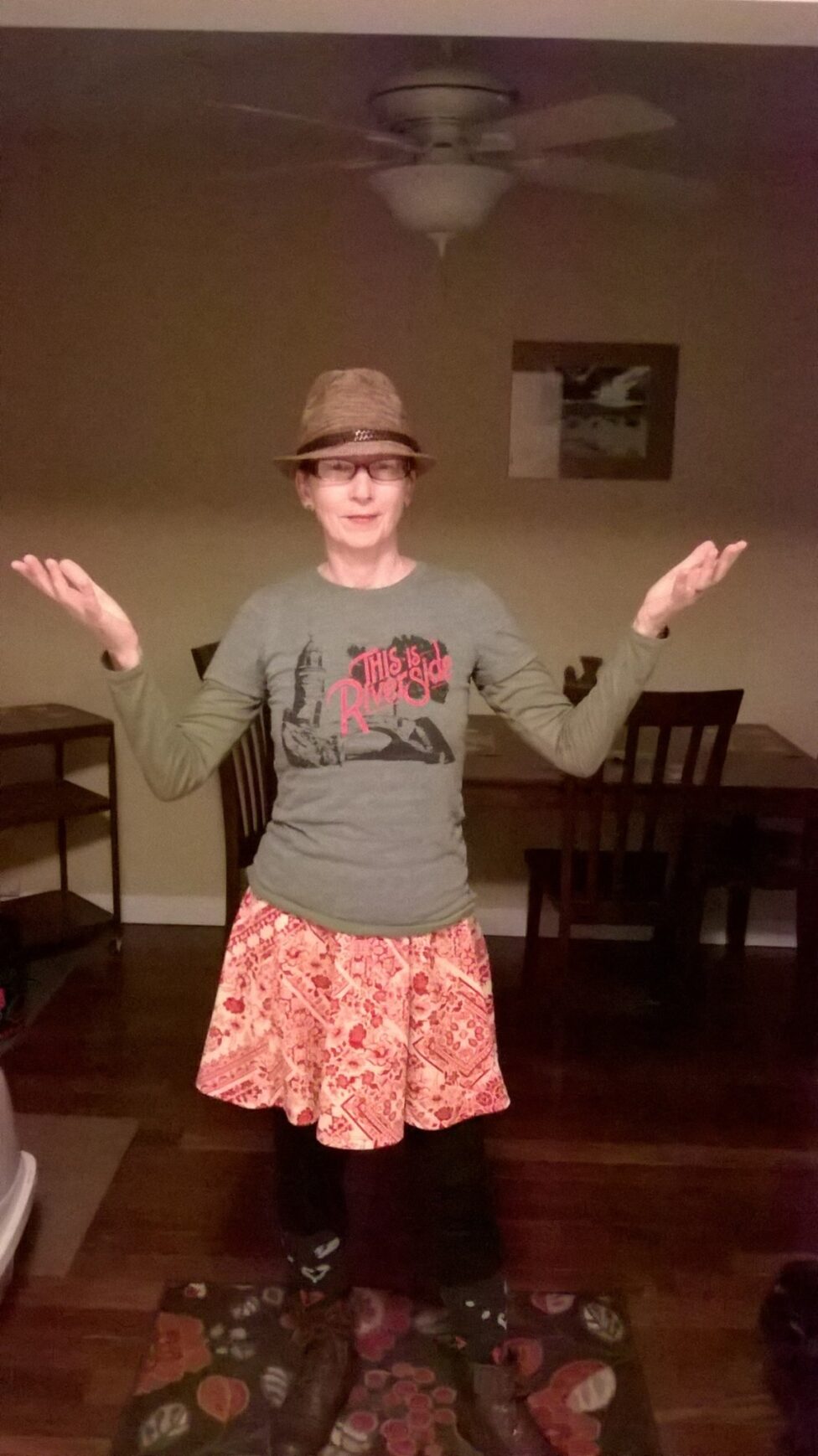 Sue faces the camera with her arms bent at the elbows and hands up in the air. She wears a grey shirt that reads “This is Riverside” and a coral patterned skirt. In the background is a brown table and chairs and a small cabinet. Photo by Sue Rogkinski.