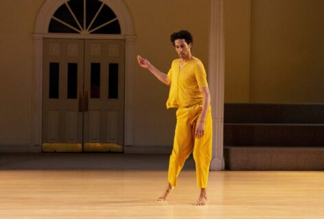 Stacy Spence is a black man alone in dance gesturing to the side as his gaze looks down to the side toward camera while in motion. He is wearing an ochre knit short sleeved shirt that buttons up the front, with ochre loose pants. Photo by Elyssa Goodman