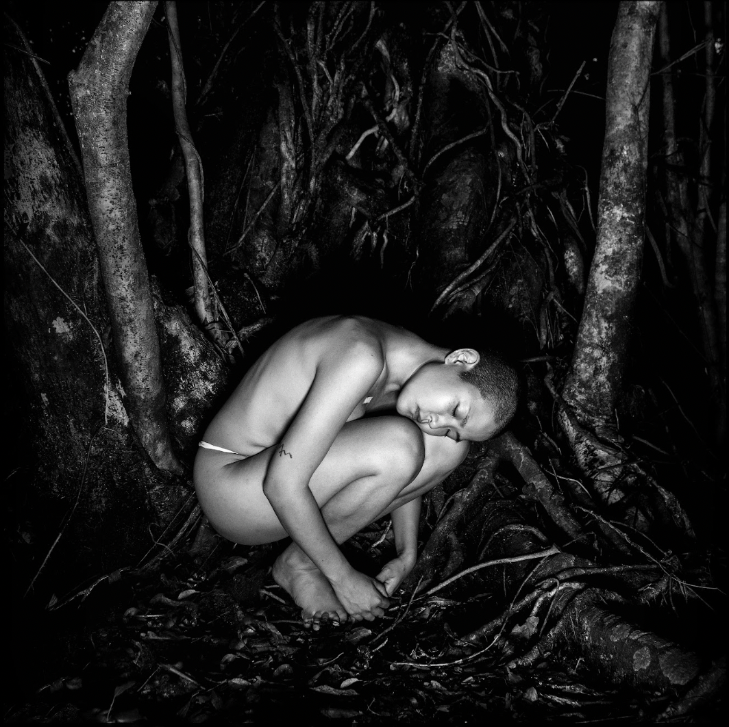 Black and white image of kyoko in a rounded, crouched position. They are not wearing clothes, and are illuminated against a dark background of trees and dirt. Photo by Andre Shizen.