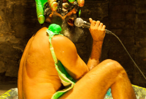 Antonio sits in a kiddie pool naked with a crown of painted dildos on his head, he holds a microphone to his mouth. Photo by Ian Douglas.